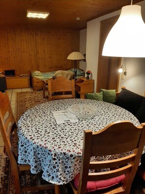 Habitació acollidora amb una taula rodona de fusta coberta per un mantell de flors, un llit de fusta amb un coixí verd i una làmpada sobre una tauleta de nit.