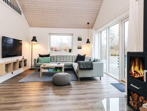 A modern living room with a grey sofa, a coffee table, a flat-screen TV, a fireplace, and a large window. - 4 Star Holiday Home in Vaeggerlose (Vaeggerlose)