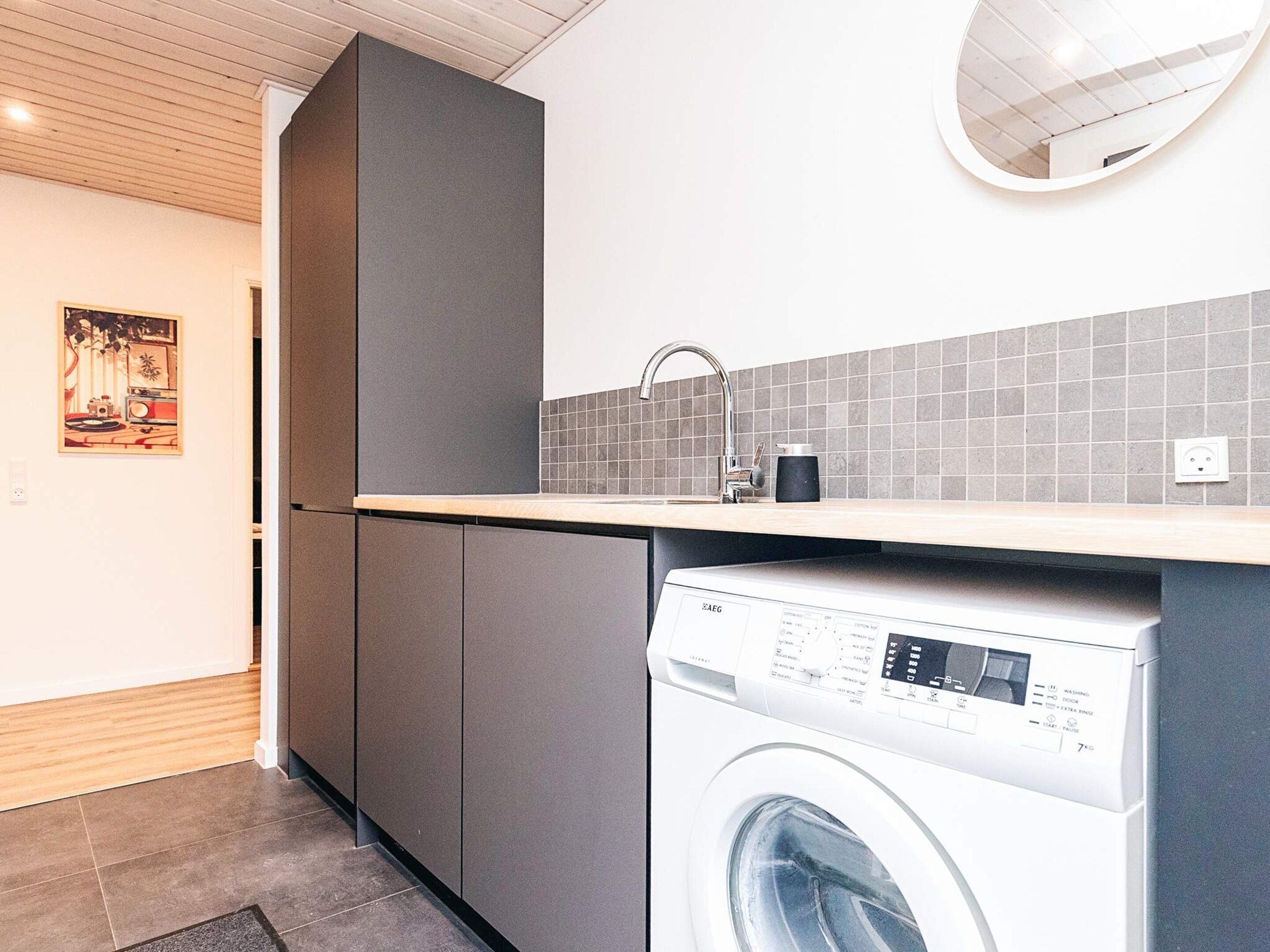 Une cuisine moderne avec des meubles gris, un lave-linge encastré et un pare-feu en carrelage.