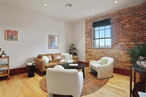 A spacious living room with a brick wall, a large window, a leather sofa, a coffee table, and a side table with a plant. - The Gisele: 4BR/3BA for 8 | Pool + Streetcar Stop (New Orleans)