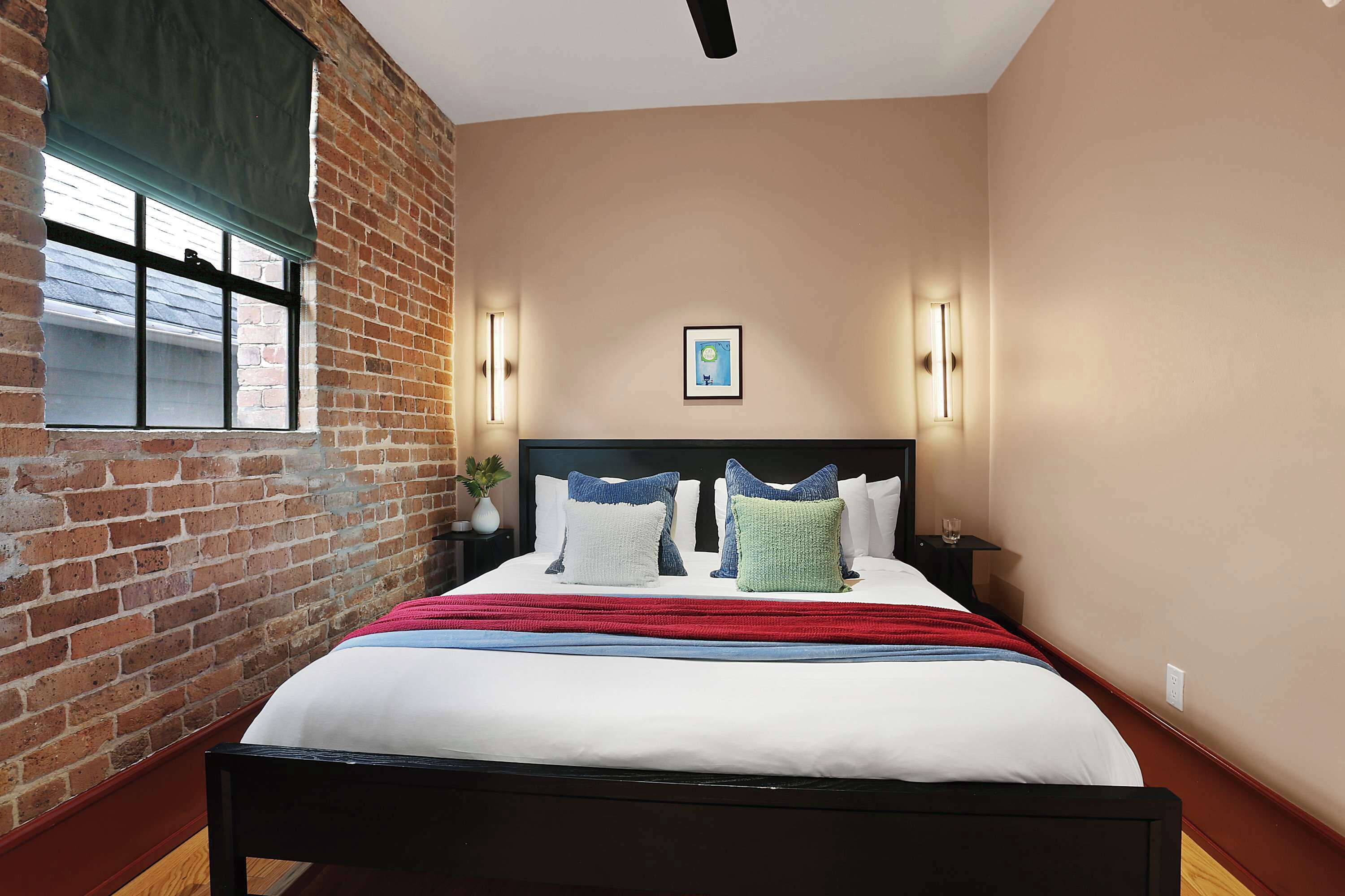 A bedroom with a brick wall, a window with blinds, a bed with multiple pillows, and wall-mounted lights.