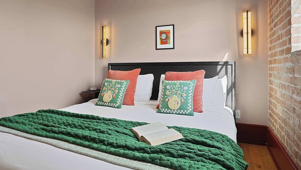 Un letto rifatto con cura, una coperta verde e due cuscini decorativi, una cornice appesa al muro e una parete in mattoni su un lato.