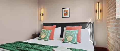 A neatly made bed with a green blanket and two decorative pillows, a framed picture on the wall, and a brick wall on one side.