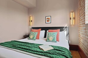 A neatly made bed with a green blanket and two decorative pillows, a framed picture on the wall, and a brick wall on one side. - The Gisele: 4BR/3BA for 8 | Pool + Streetcar Stop (New Orleans)