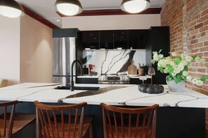 Une cuisine moderne avec un îlot central, des meubles de couleur noire, des appareils électroménagers en acier inoxydable et un mur en briques.