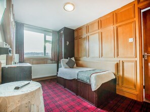 A room with a bed, a small table, a radiator, a window with curtains, and wooden paneling. - Standard Single Room (Gateshead)