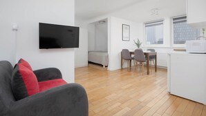 Een modern appartement met een zithoek met een grijze bank voorzien van rode kussens, een flatscreen televisie aan de muur, een eethoek met een tafel en stoelen, en een kitchenette met een magnetron.