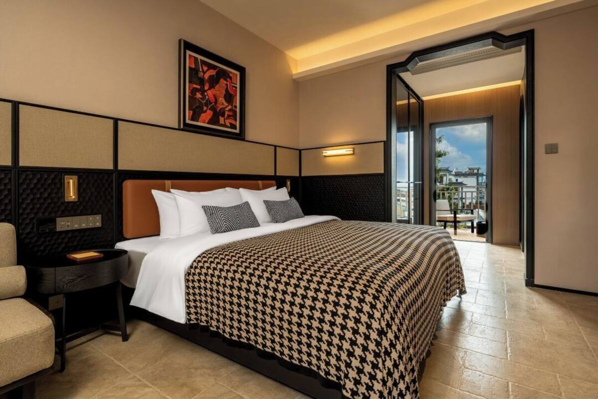 Ein Hotelzimmer mit einem großen Bett, einem Nachttisch, einem Sessel und Blick ins Freie.