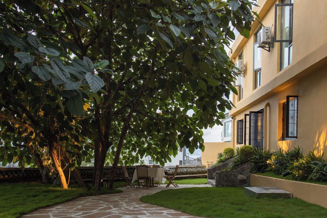 Um pátio com caminho de pedra, área de refeições com mesas e cadeiras, e um edifício com janelas amplas e fachada amarela.