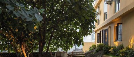 Um pátio com caminho de pedra, área de refeições com mesas e cadeiras, e um edifício com janelas amplas e fachada amarela.