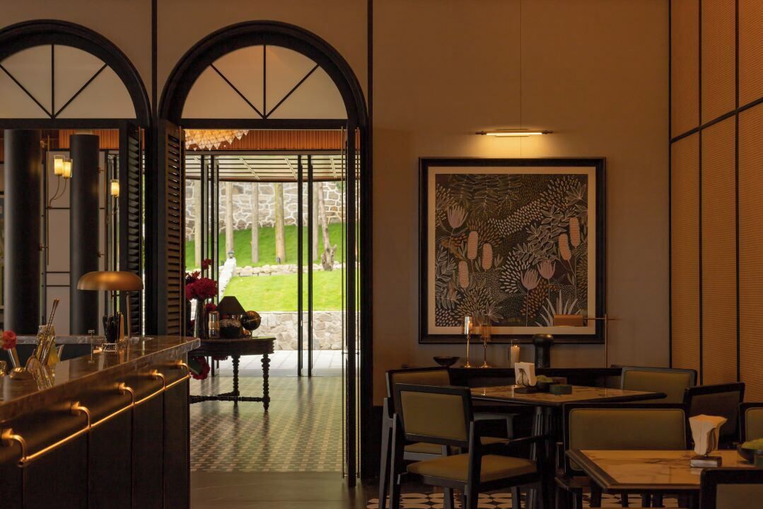 A restaurant with a bar area, dining tables, and chairs. There is a large framed artwork on the wall and a view of a garden through the open door.