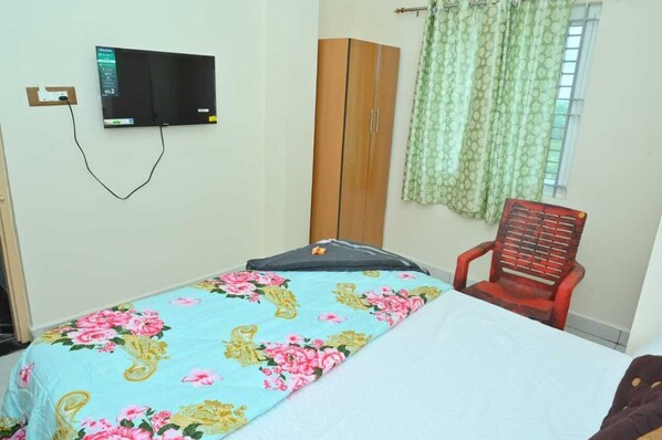 A bedroom with a bed, a television mounted on the wall, a wardrobe, and a window with curtains. - Radha Krishna Heritage (Hospet)