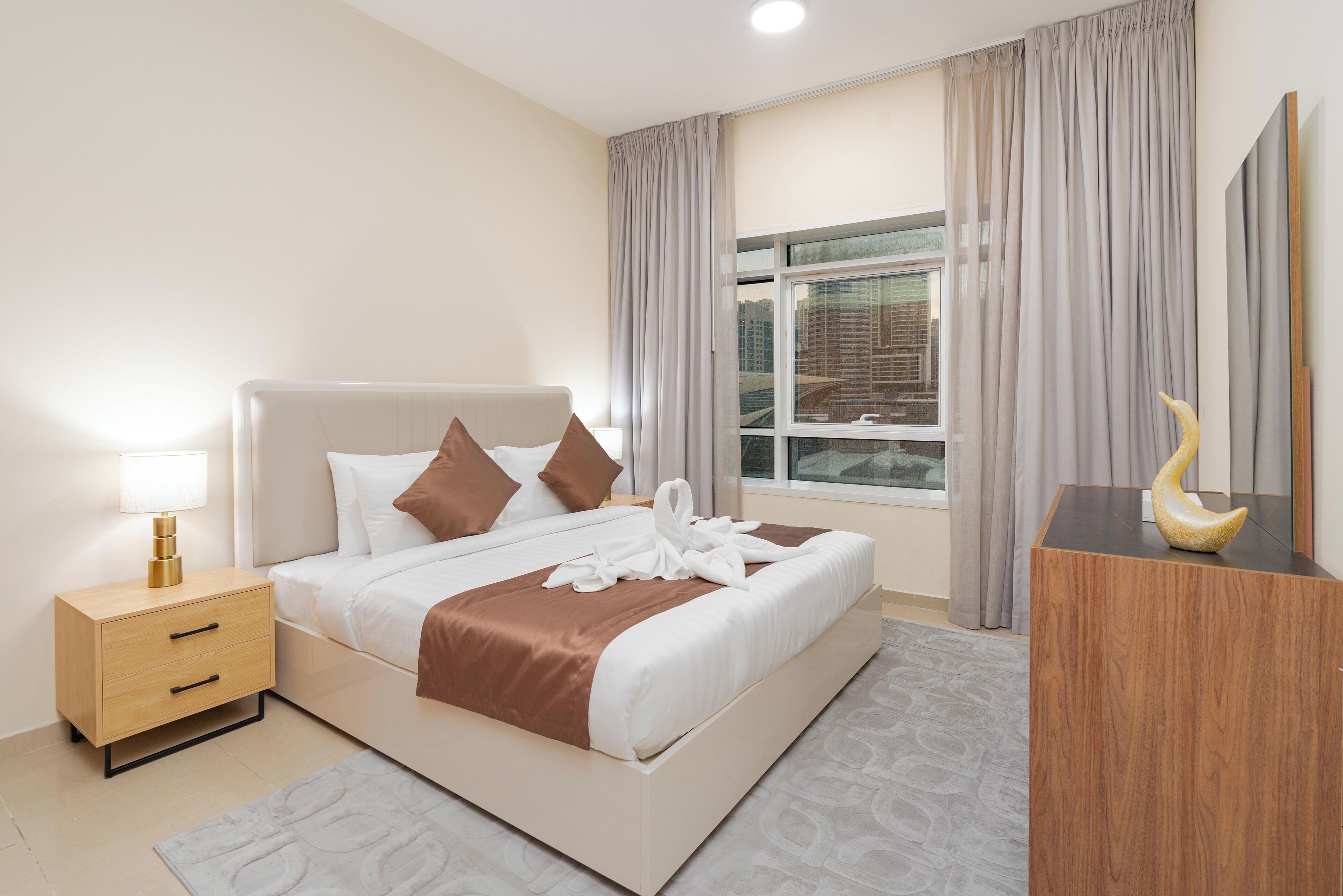 Ein modernes Hotelzimmer mit einem großen Bett, einem Nachttisch mit Lampe, einem Holzbeistelltisch, einem Spiegel und Blick auf die Stadt.