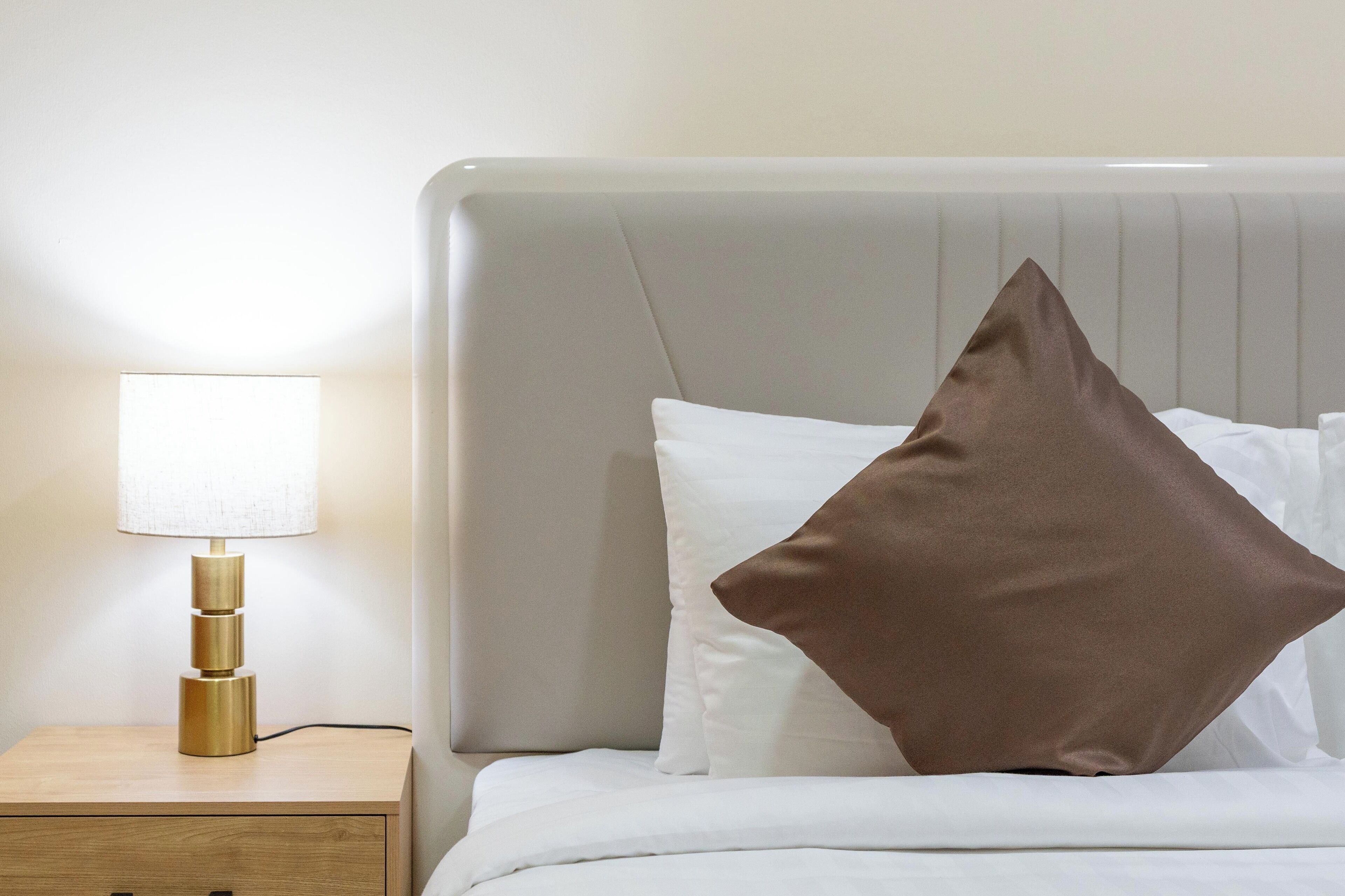 A neatly made bed with white bedding and a single brown pillow, a bedside table with a lamp, and a light-colored headboard.