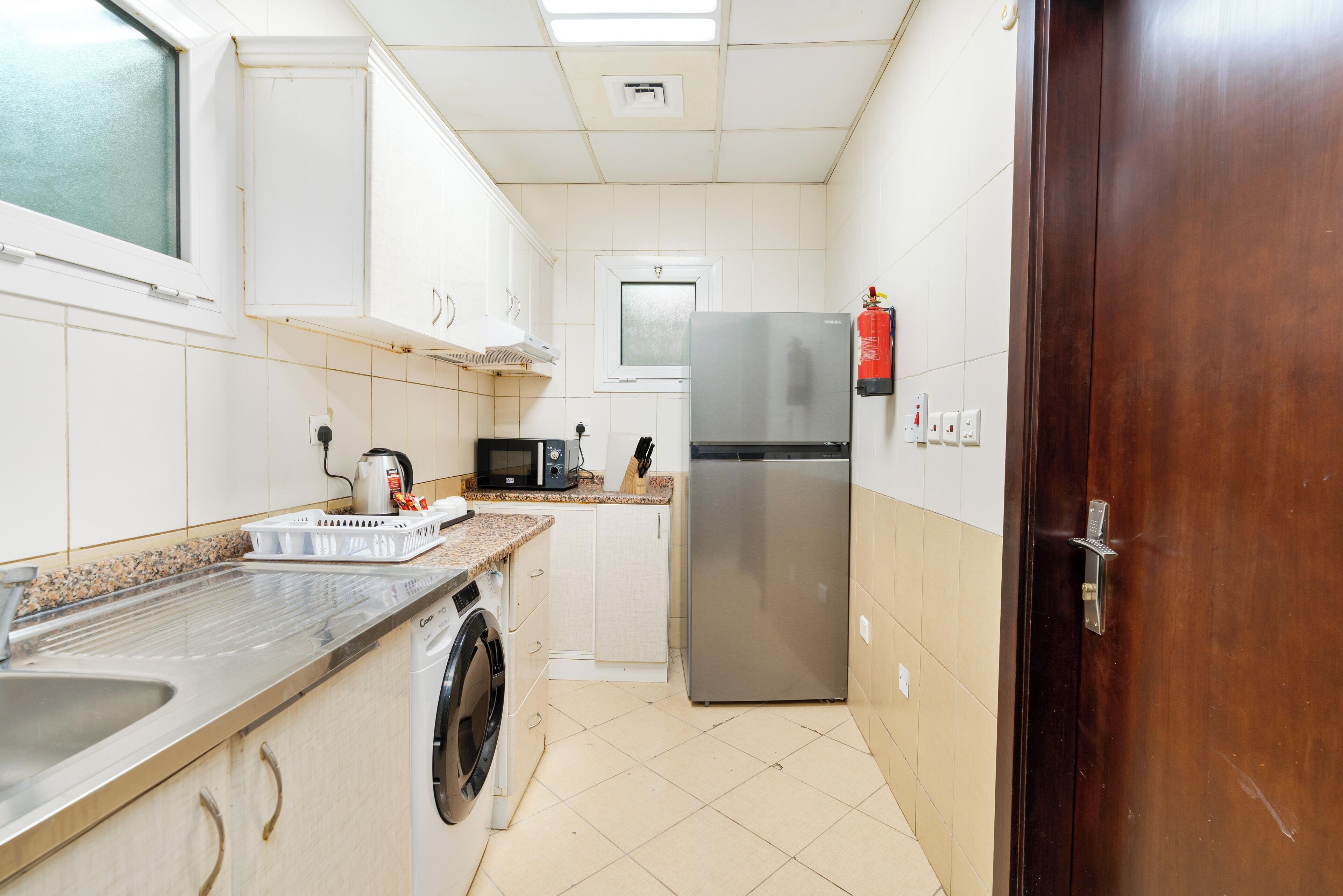 A compact kitchen with a stainless steel refrigerator, a washing machine, a microwave, and a sink.