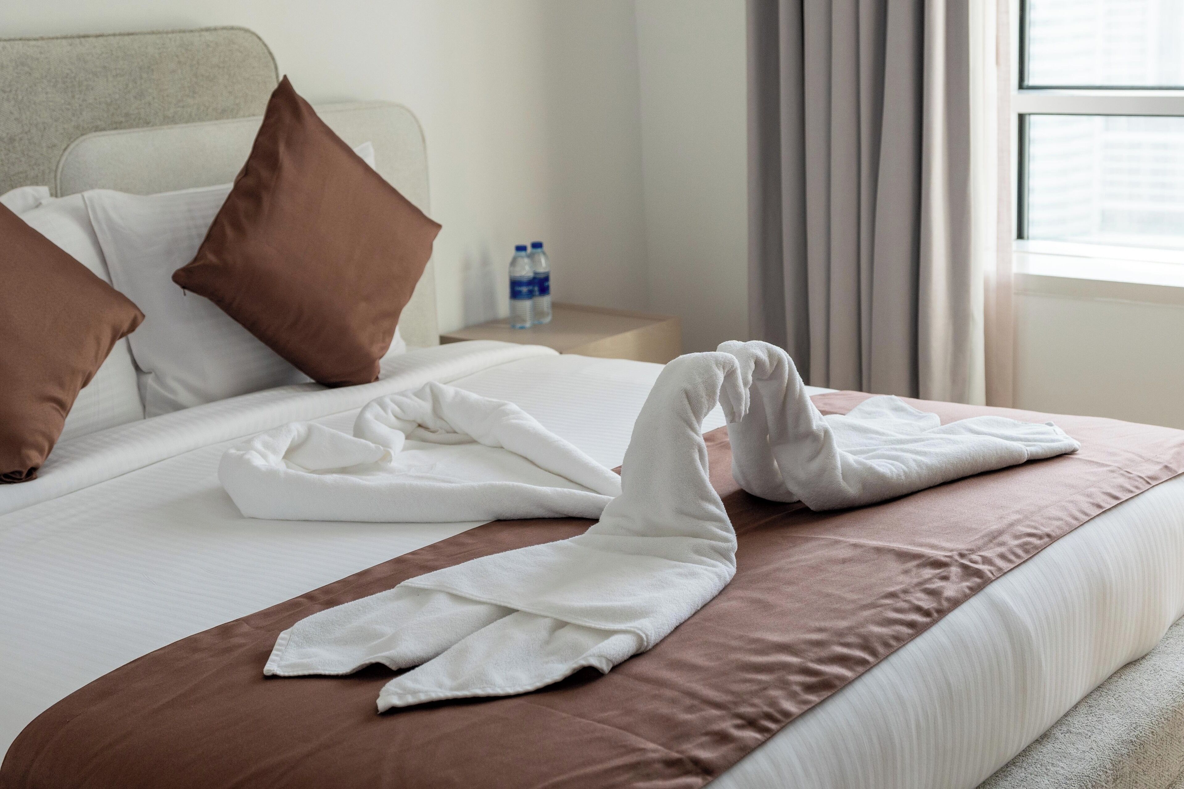A neatly made bed with brown and white pillows, a water bottle on a nightstand, and a window with curtains.