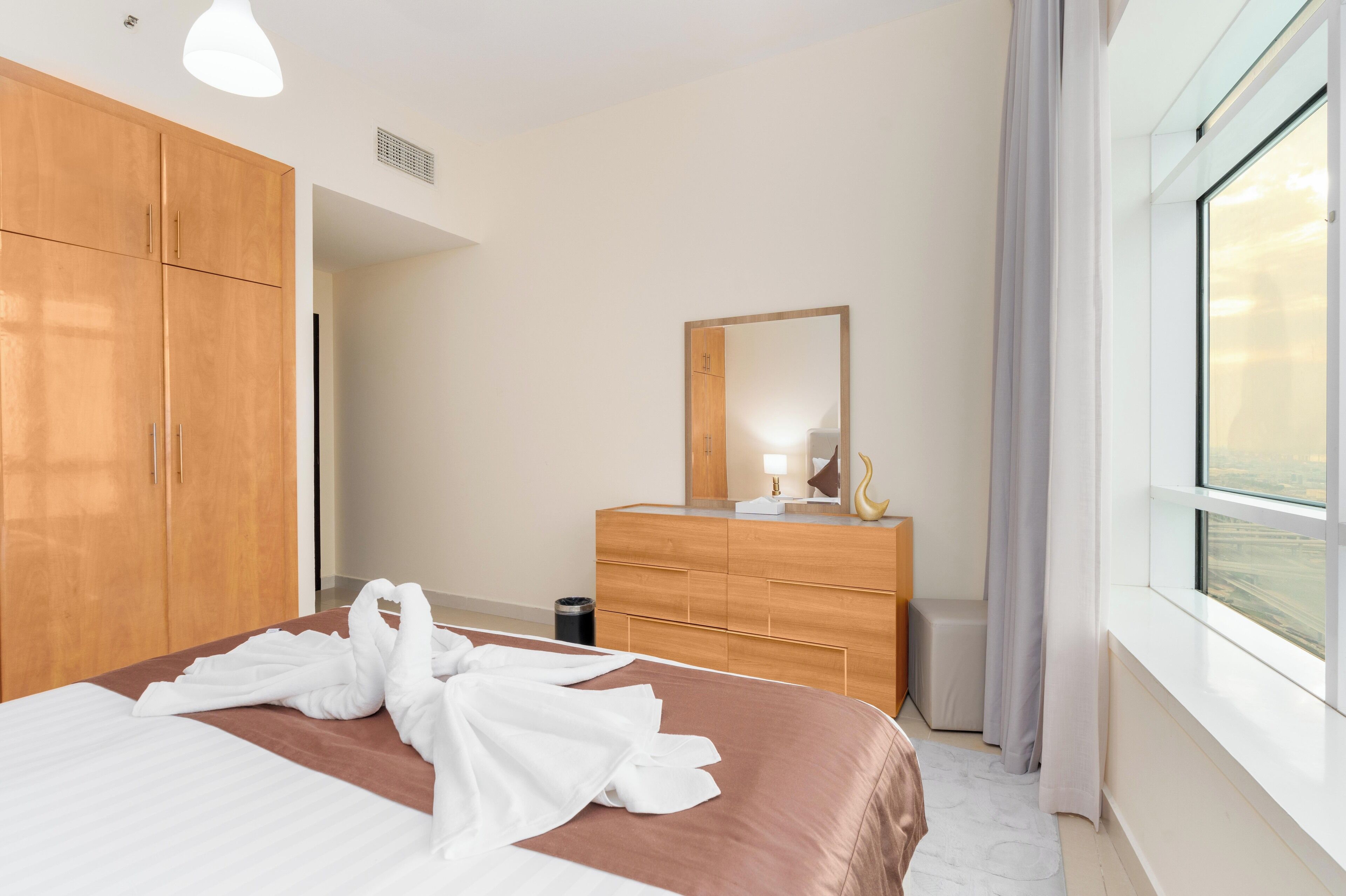 A hotel room with a bed, wooden wardrobe, a dresser, a mirror, and a window with curtains.