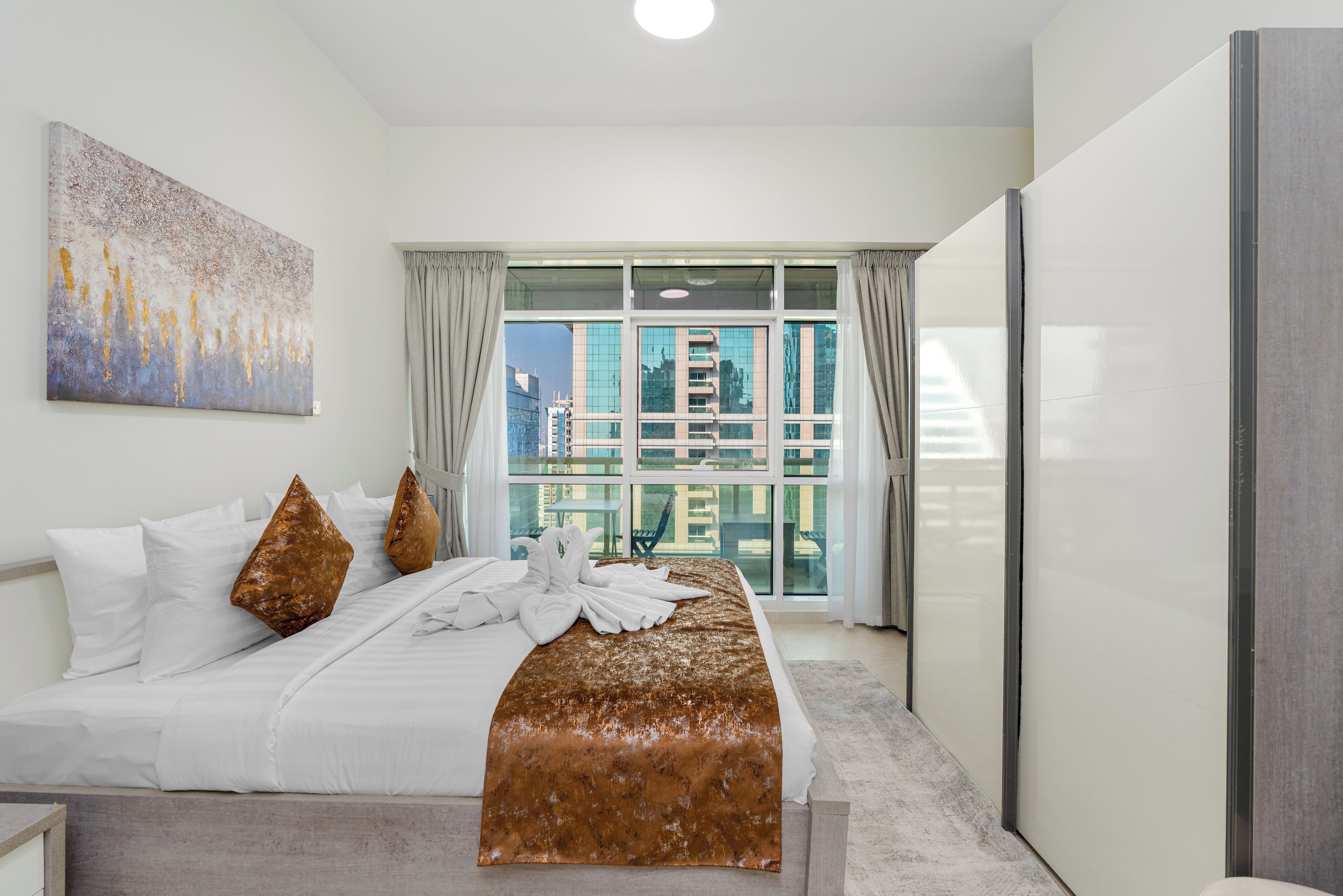 A hotel room with a large bed, a view of buildings through the window, and a painting on the wall.