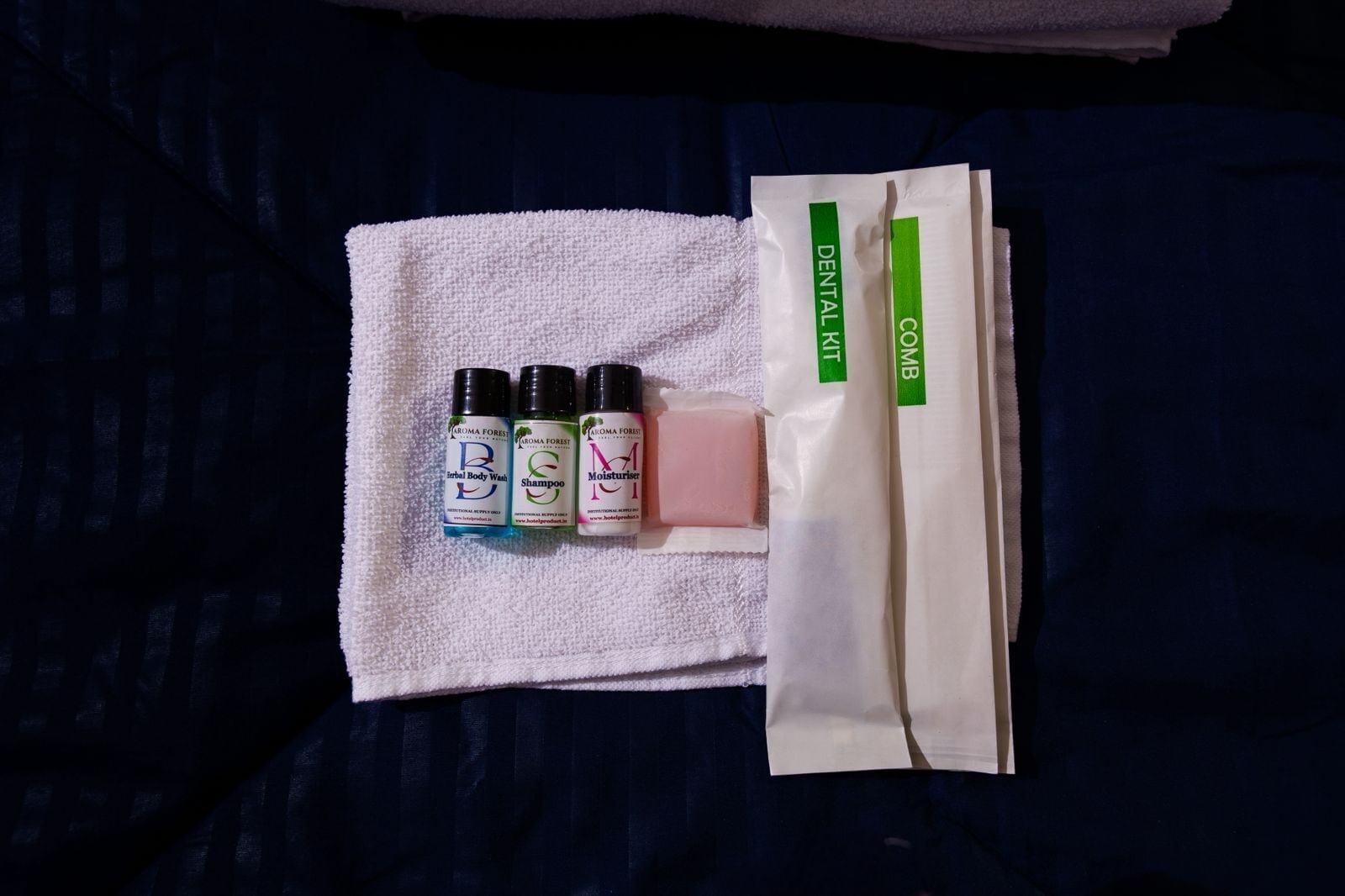 A white towel with three bottles of body wash, shampoo, and moisturizer, and a dental kit on a dark blue fabric.