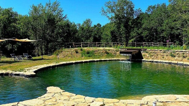 Una piscina con borde de piedra, un puente de madera y una zona sombreada con sillas.
