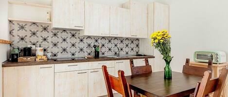A kitchen with light wooden cabinets, a tiled backsplash, a dining table with chairs, and a small green vase with yellow flowers.