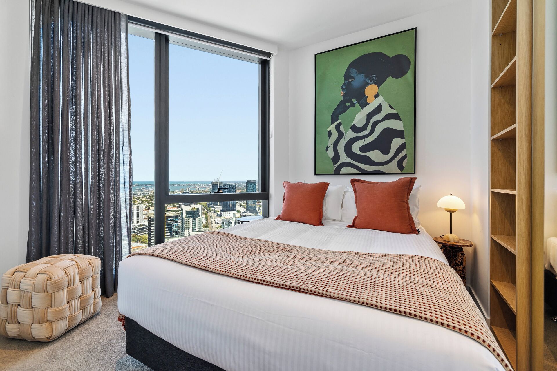 Quarto de hotel com uma cama grande, vista da cidade, luminária moderna e uma pintura abstrata colorida na parede.