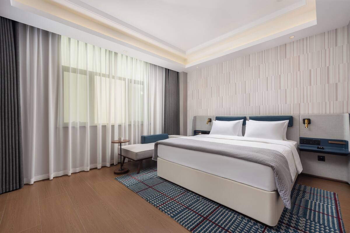 Ein modernes Hotelzimmer mit einem großen Bett, einem Nachttisch, einem Beistelltisch und einem Fenster mit leichtem Vorhang.