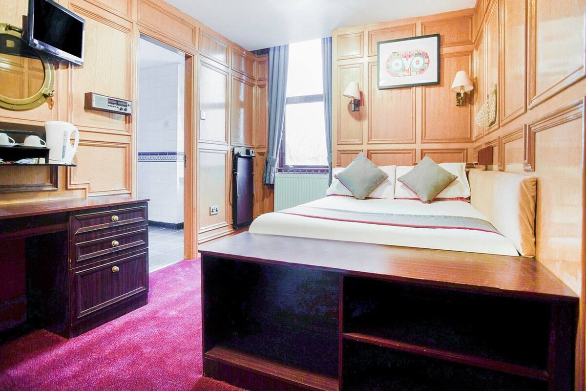 A hotel room with wooden paneling, a bed with pillows, a TV mounted on the wall, a desk with drawers, and a radiator.