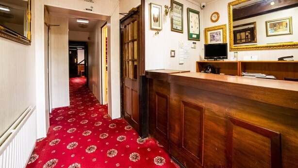 Un hall d’hôtel avec un tapis rouge, un comptoir d’accueil en bois et des tableaux encadrés accrochés au mur.