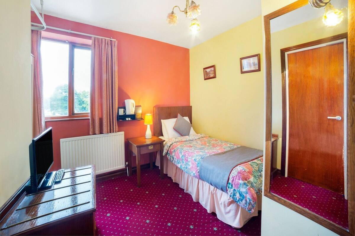 A hotel room with a bed, a desk, a chair, a radiator, a window with curtains, a lamp, and a door.