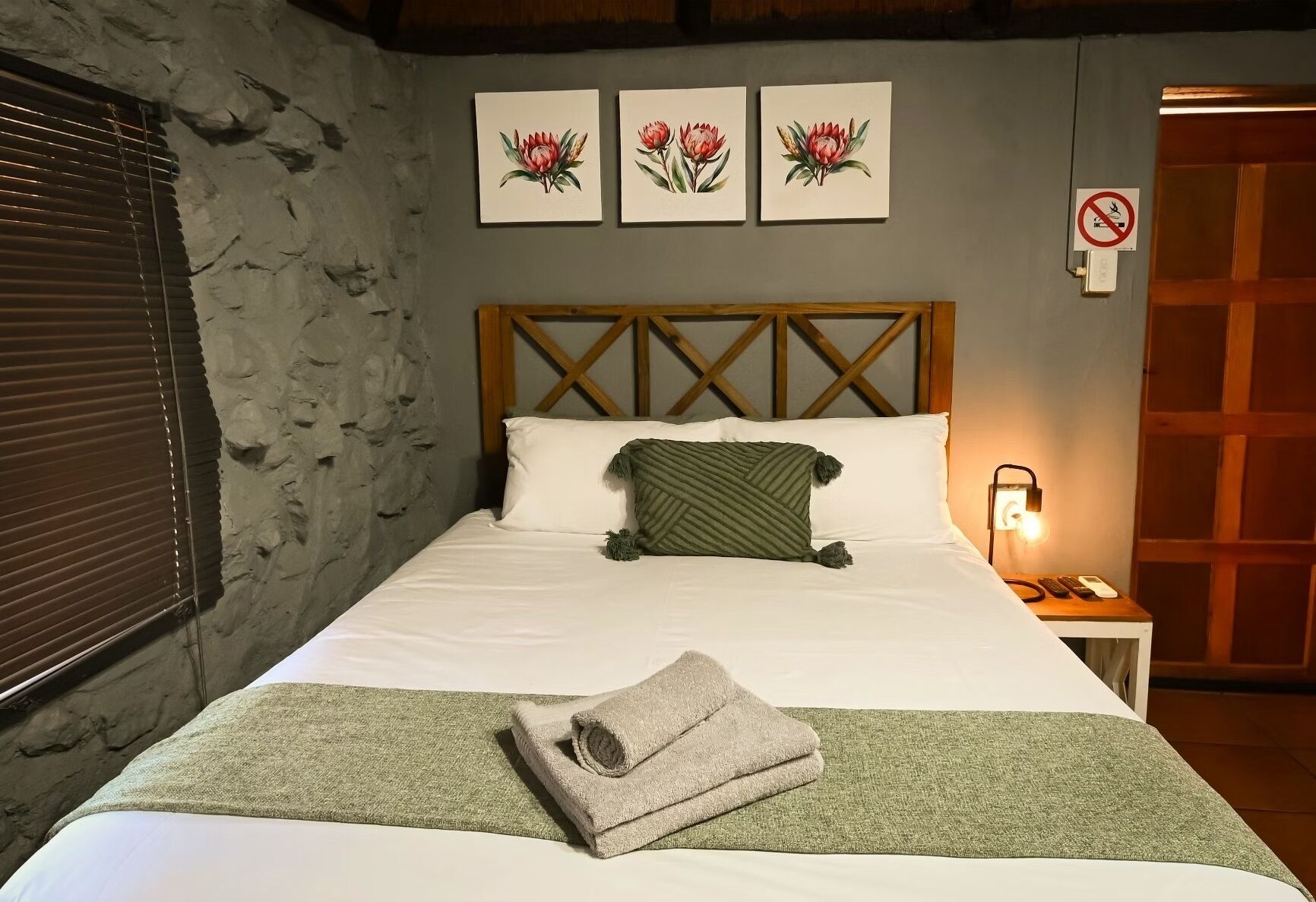 Ein ordentlich bezogenes Bett mit grüner Decke und gerolltem Handtuch, ein hölzernes Kopfteil sowie drei gerahmte Blumenbilder an der Wand.