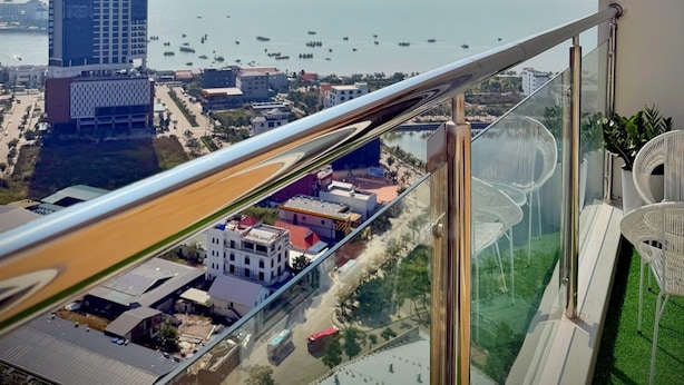 Un balcon doté d’une balustrade en verre donnant sur un paysage urbain, avec notamment un immeuble de grande hauteur et une étendue d’eau où naviguent des bateaux.