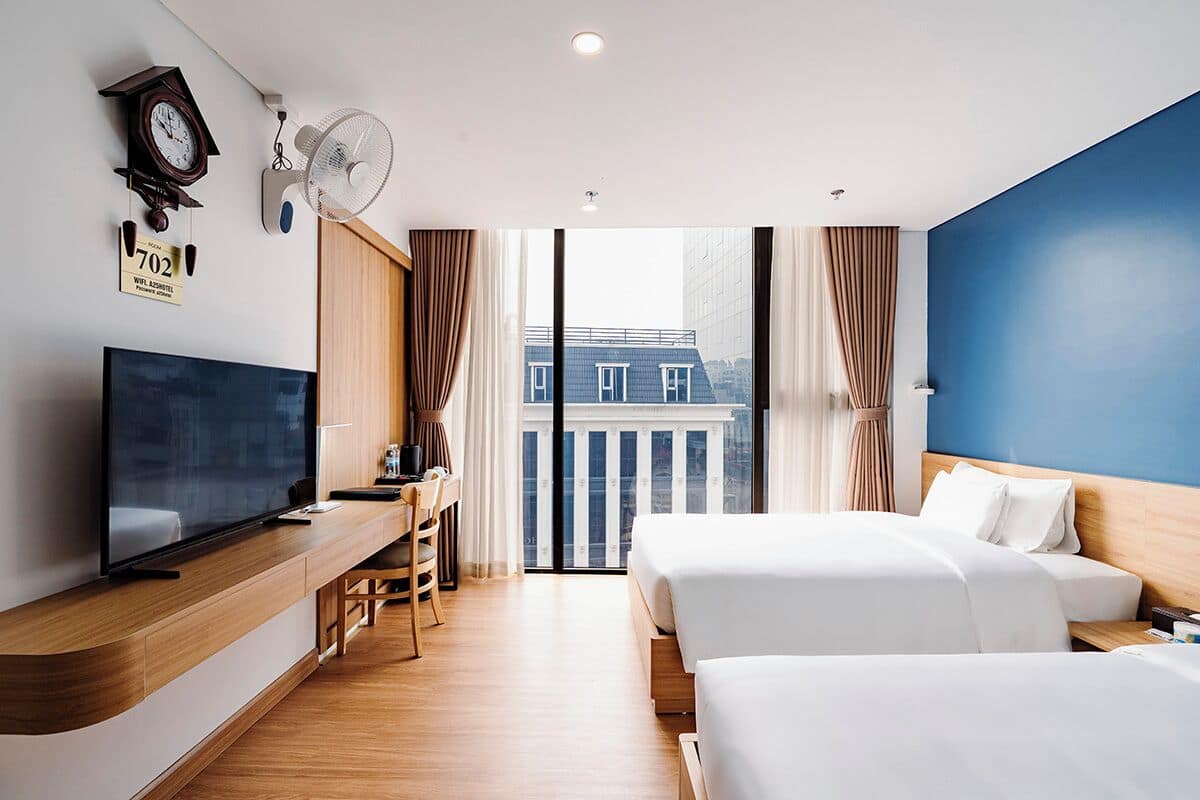 Een hotelkamer met een televisie, bureau, twee bedden, klok, ventilator en uitzicht op een gebouw.