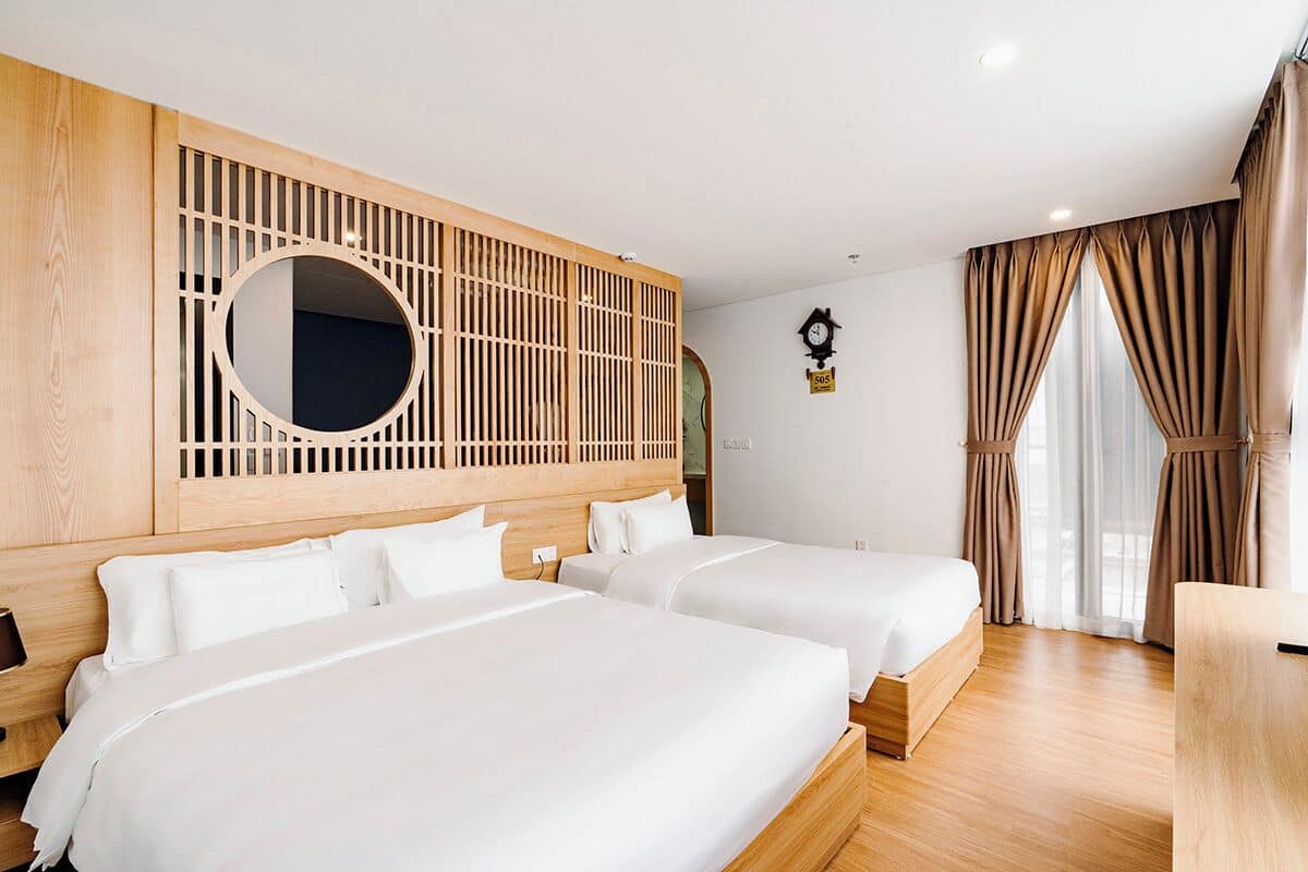 Een hotelkamer met twee bedden, een houten hoofdbord, een ronde spiegel, een klok en uitzicht op het omliggende landschap door grote ramen.