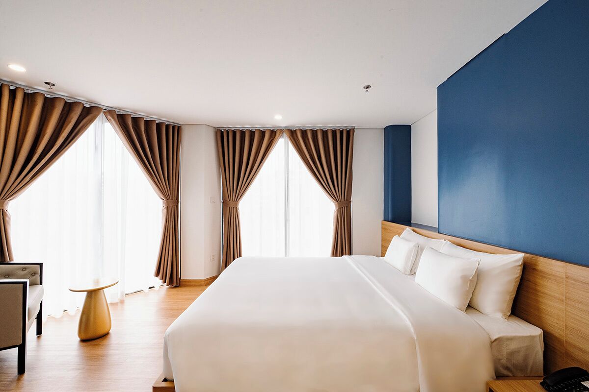 Habitación de hotel con una cama grande, cabecero de madera, una mesita redonda y cortinas en las ventanas.