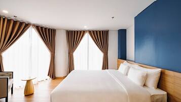 Habitación de hotel con una cama grande, cabecero de madera, una mesita redonda y cortinas en las ventanas.