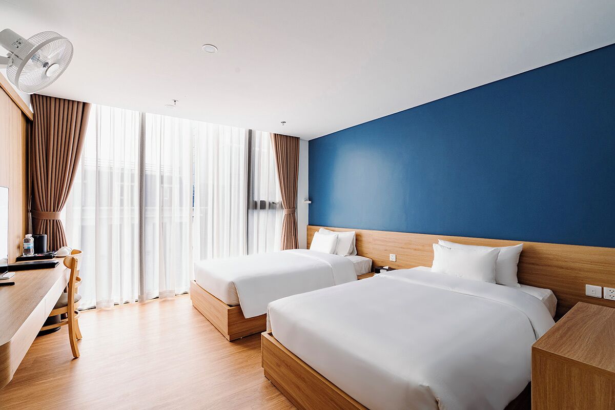 Une chambre d’hôtel avec deux lits, un bureau, une chaise, un ventilateur et une vue sur l’extérieur.