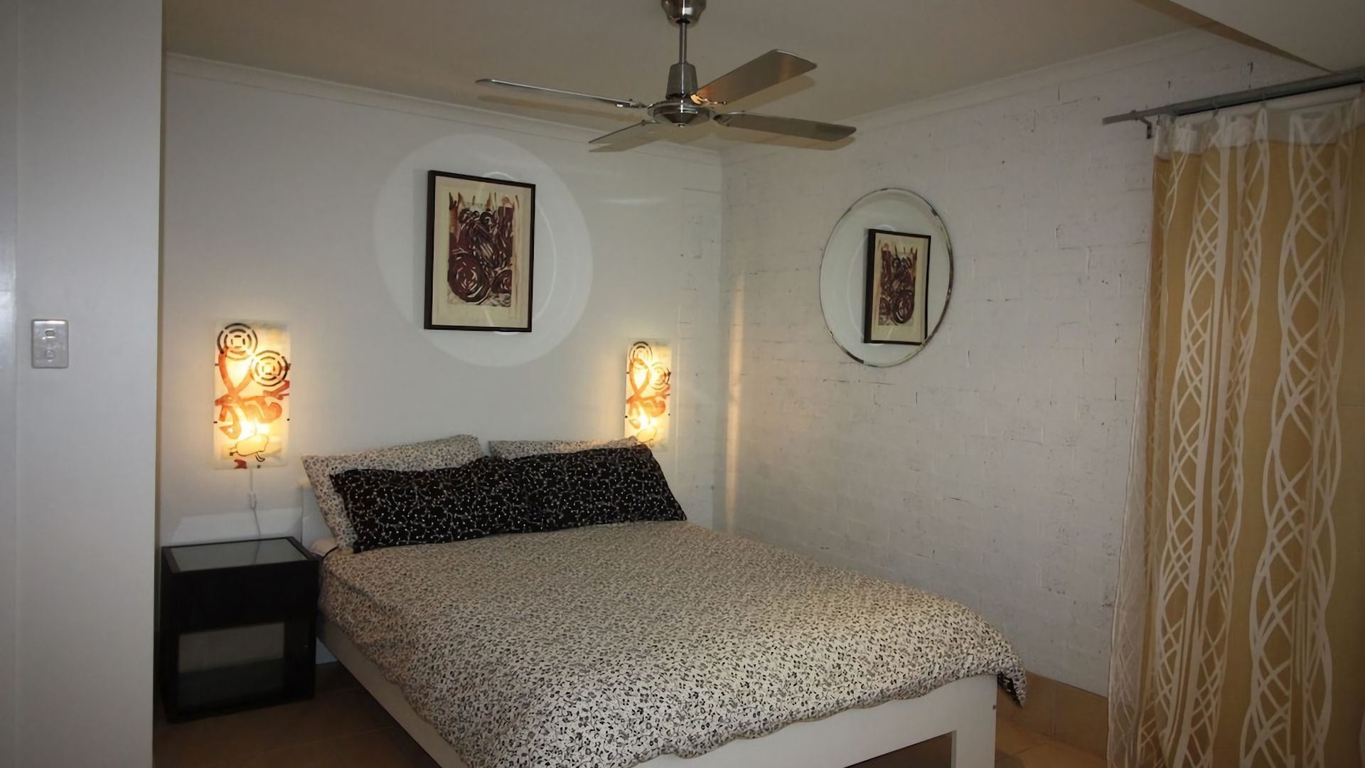 A bedroom with a bed, a ceiling fan, two wall-mounted light fixtures, and a window with a curtain.