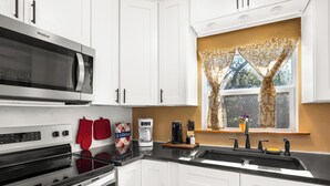 A kitchen with white cabinets, a stainless steel microwave, a coffee maker, and a window with floral curtains. - Fabulous Hill Country Treehouse • Relaxing Stay with All Amenities (Lakehills)