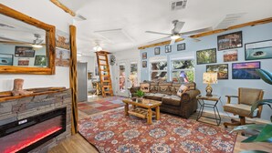 A cozy living room with a fireplace, a ceiling fan, and a large rug. - Fabulous Hill Country Treehouse • Relaxing Stay with All Amenities (Lakehills)