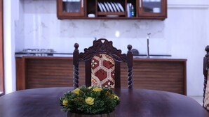 Une table à manger agrémentée d’un centre de table floral, une chaise en bois avec un dossier à motifs, et un coin cuisine avec des placards en bois et une cuisinière en arrière-plan.
