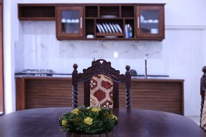 A dining table with a floral centerpiece, a wooden chair with a patterned backrest, and a kitchen area with wooden cabinets and a stove in the background. - Luxury Type 3-bedroom villa in perfect Unawatuna with cool AC comfort (Unawatuna)