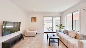 Uma sala de estar moderna com uma televisão de tela plana, um sofá bege, uma poltrona branca e uma mesa de centro. Há uma porta de vidro deslizante que dá para uma área de varanda externa.