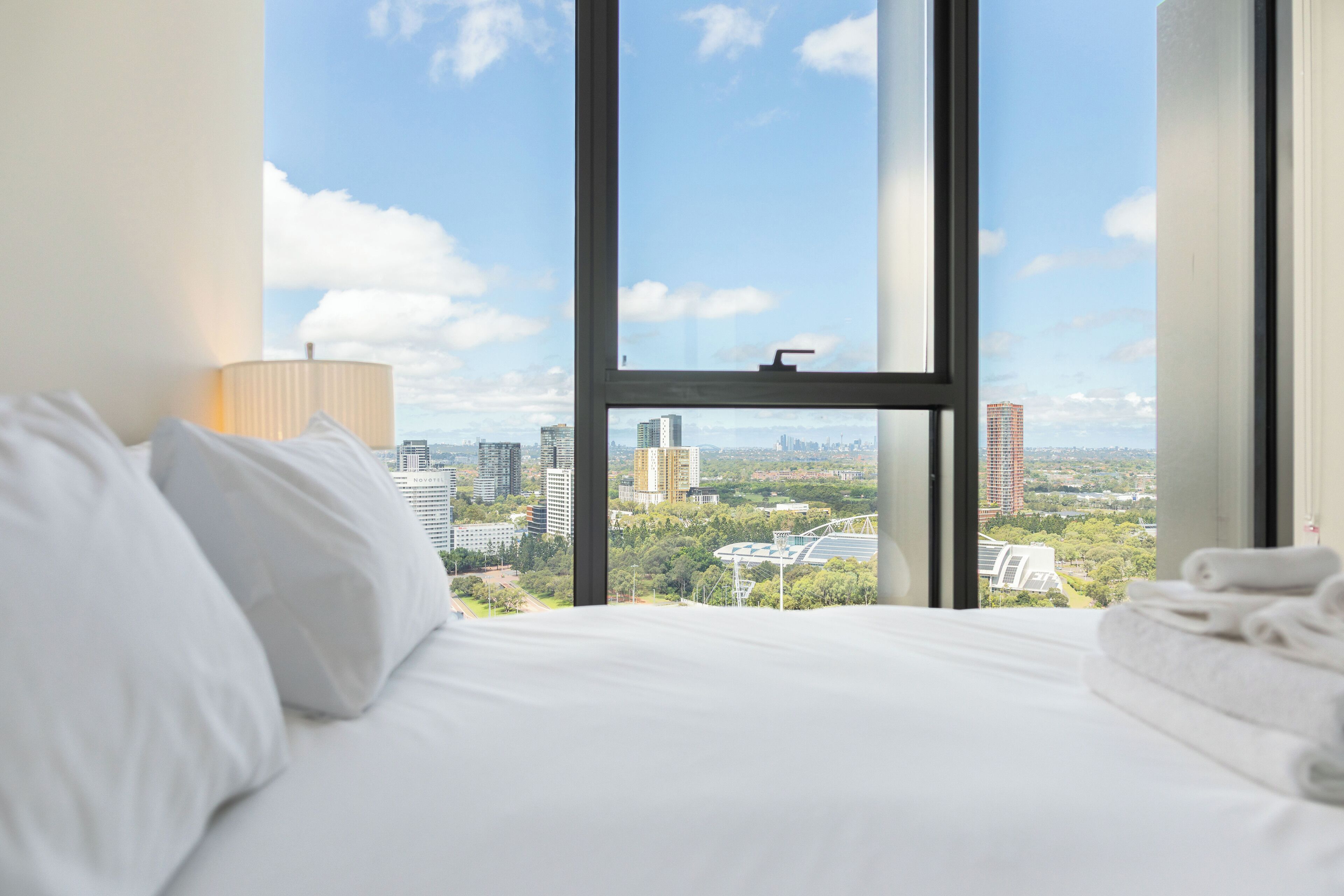 A hotel room with a large window offering a city view, a neatly made bed, and a lamp on the side.