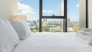 Quarto de hotel com uma grande janela com vista para a cidade, uma cama bem arrumada e um abajur ao lado.