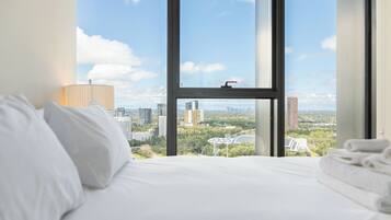 A hotel room with a large window offering a city view, a neatly made bed, and a lamp on the side.