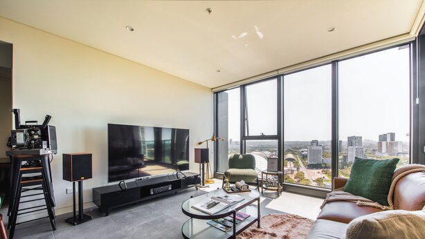 Uma sala de estar moderna com uma televisão grande, assentos confortáveis, uma mesa de centro de vidro e vista para a cidade por grandes janelas.