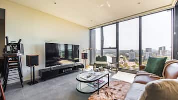 A modern living room with a large TV, comfortable seating, a glass coffee table, and a city view through large windows.