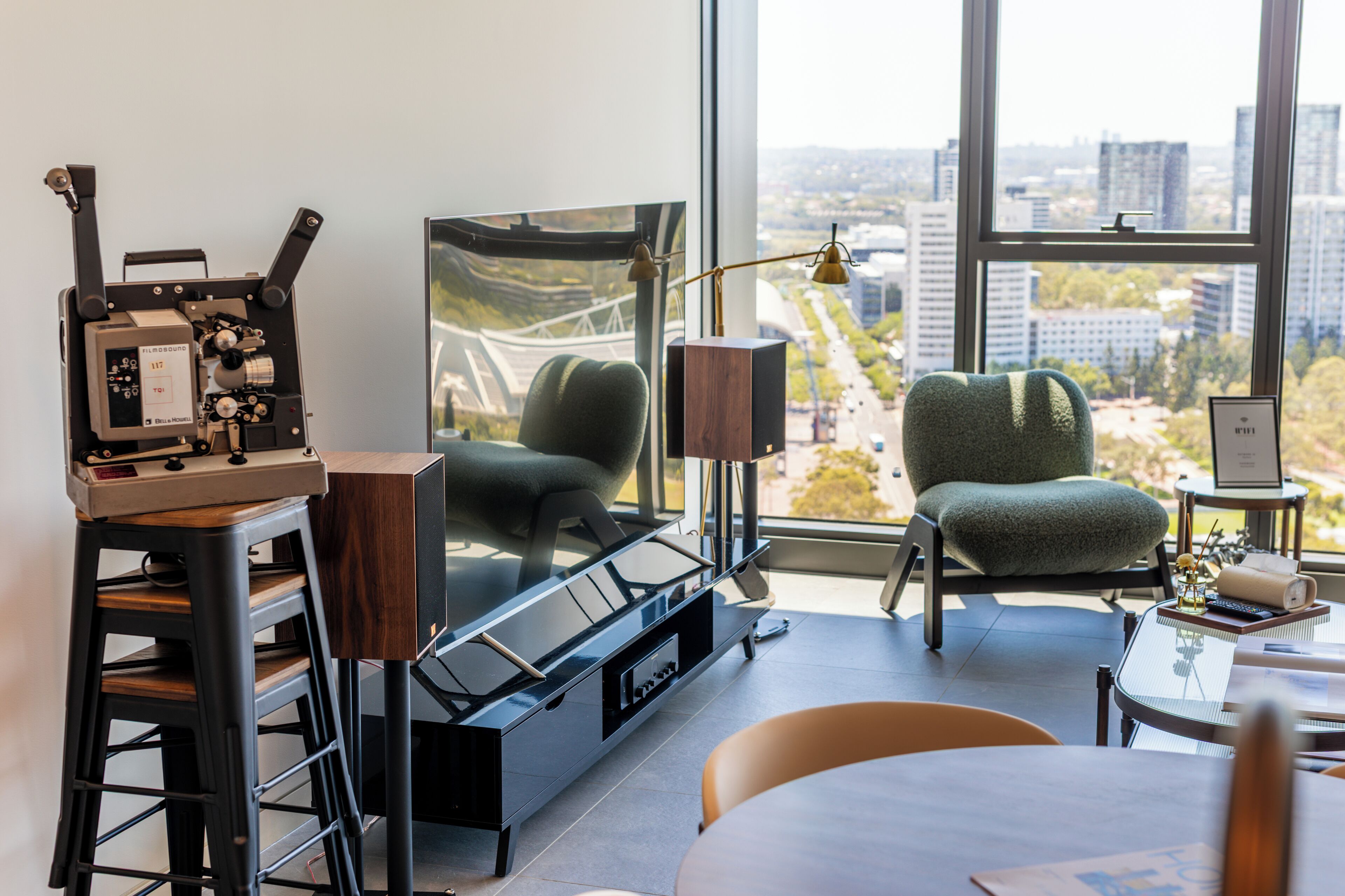 A modern room with a large window, a vintage film camera, a green armchair, and a table with a lamp.