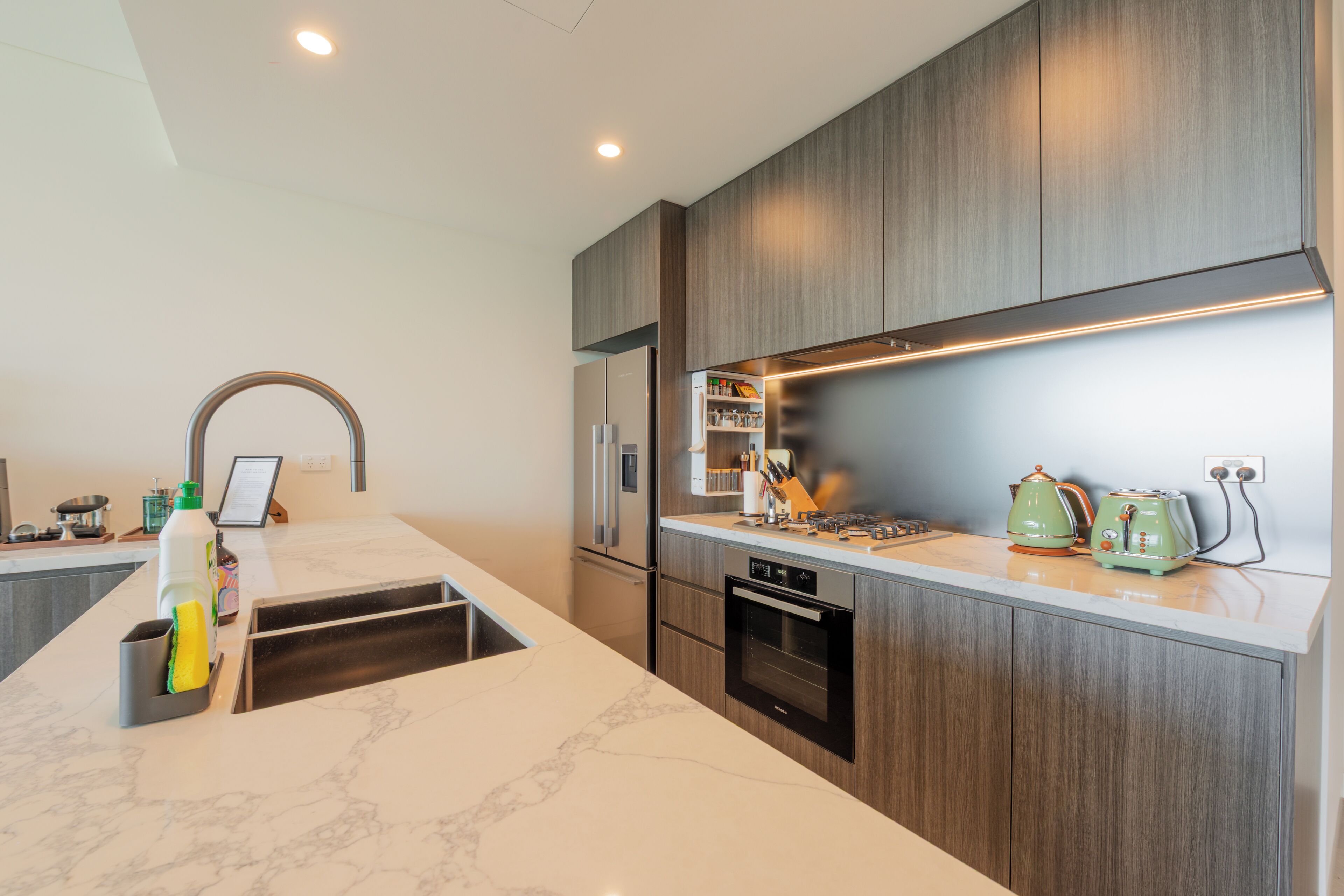 A modern kitchen with a marble countertop, wooden cabinets, a built-in oven, and a stainless steel refrigerator.